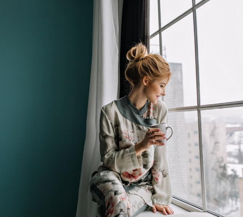 young-blonde-woman-drinking-tea-coffee-looking-through-big-window-happy-good-morning-home-wearing-silk-pajamas-with-flowers-turquoise-wall (1)
